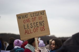 women holding up sign that says "what lessens one of us lessens all of us"