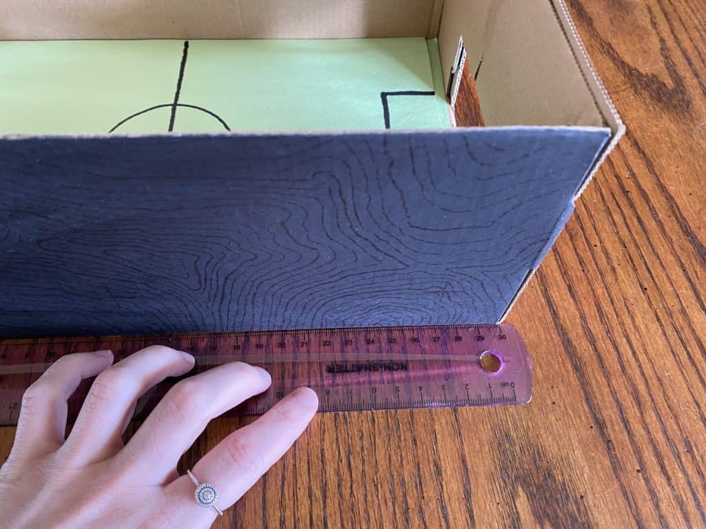 A hand holding a ruler against a black box on a wooden table.