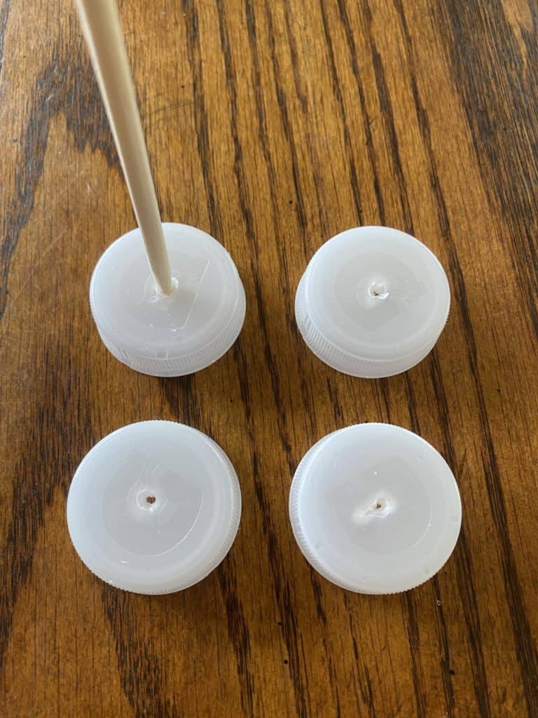 Bottle caps with holes pocked into the tops laying a table, with someone stabbing a hole into a bottle cap with a wooden stick in the left corner.