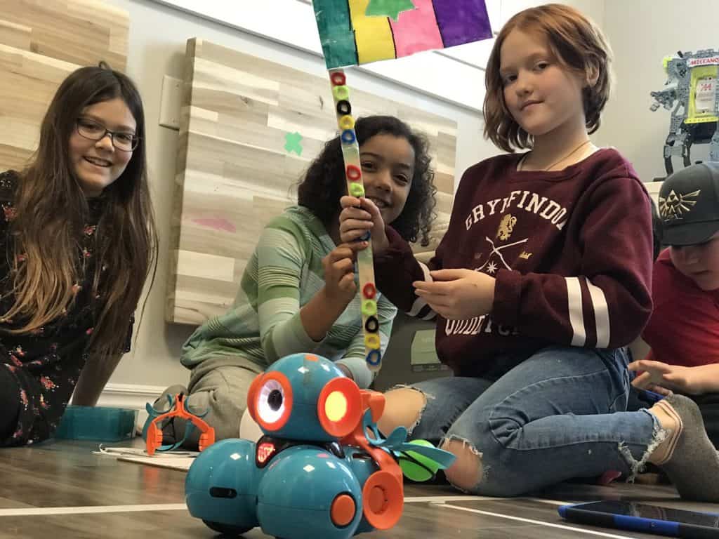 Students sitting on the floor with Dash and Dot robots in the Lindsay Makerspace, holding up a handmade flag.