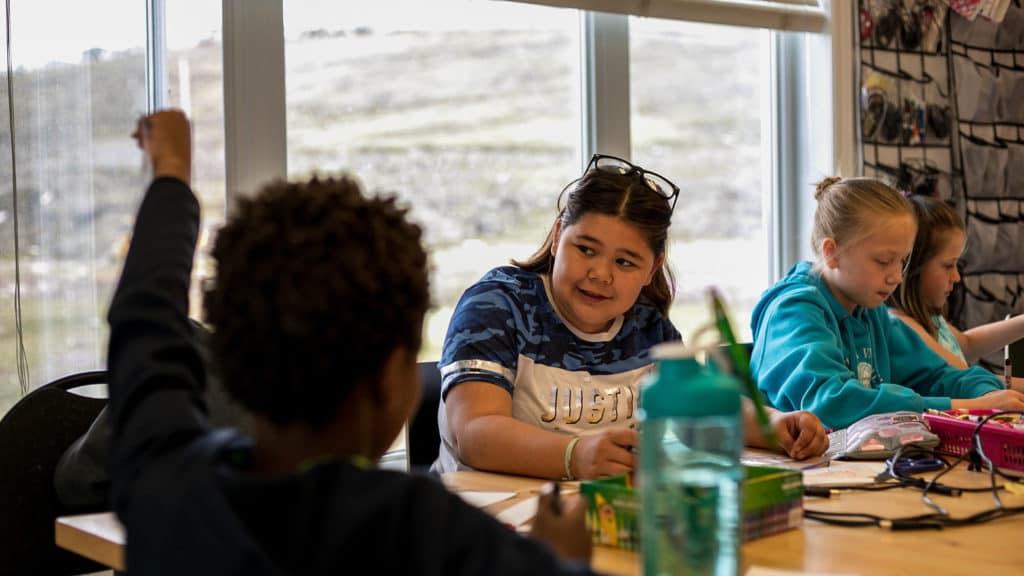 Kids looking at each other while sitting at a table with a big window behind them. One is facing the camera, the other has their back to the camera. They're learning and have writing tools in their hands.