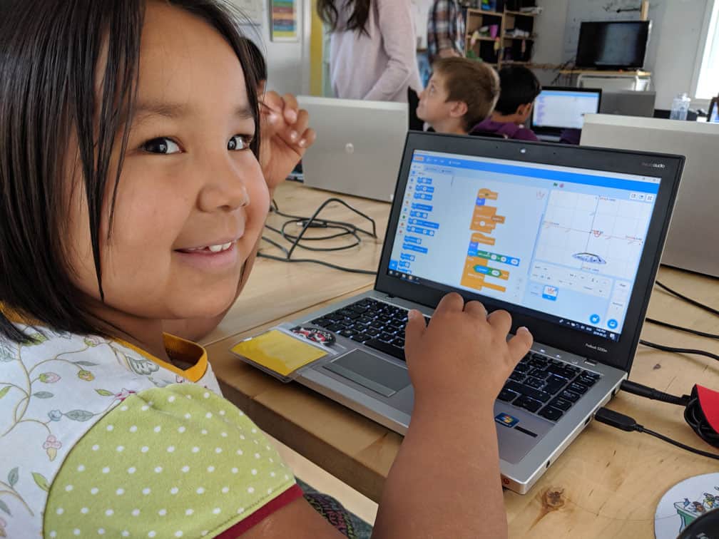 The camera is behind the learner and the learner is turned around smiling. The learner is working on Scratch on their computer.