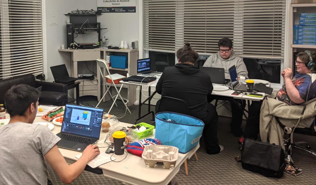 Nunavut Game Jam, 4 people are sitting down at 2 tables working on their games.